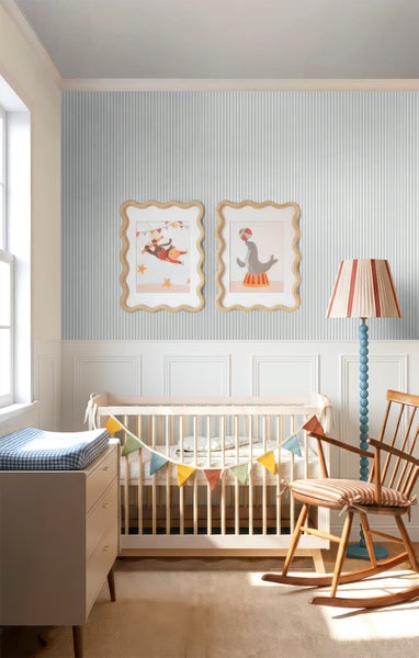 Helles Kinderzimmer mit gestreifter Tapete, weißer Wandtäfelung, Holz-Babybett, Schaukelstuhl und bunten Wandbildern im Zirkus-Stil.
