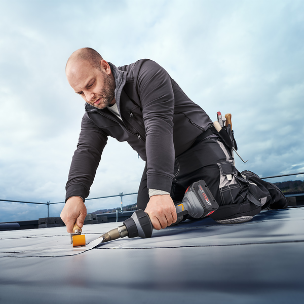 Handwerker bei der professionellen Dachabdichtung mit einem Akku Heißluftgebläse und einer Andruckrolle auf einem Flachdach.
