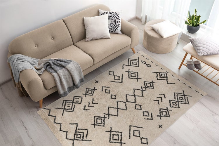 Wohnzimmer mit beige Sofa, Pouf und Teppich mit schwarzen geometrischen Mustern und Fransen auf hellem Boden.