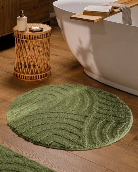 Runder grüner Badteppich mit Wellenstruktur auf Holzfußboden neben einer weißen Badewanne und einem Beistelltisch aus Rattan.