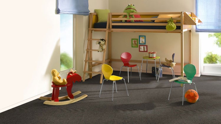 Dunkelgraue Teppichfliesen in einem hellen Kinderzimmer mit Hochbett aus Holz, bunten Stühlen und Spielzeug.