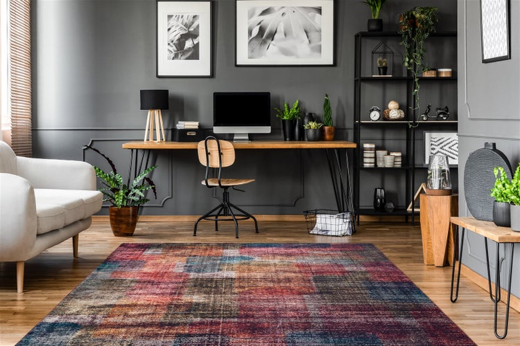 Moderner, organisierter Arbeitsbereich mit buntem Patchwork-Teppich, Holzschreibtisch, schwarzem Metallregal und hellem Sofa vor einer Wand mit Zierleisten.