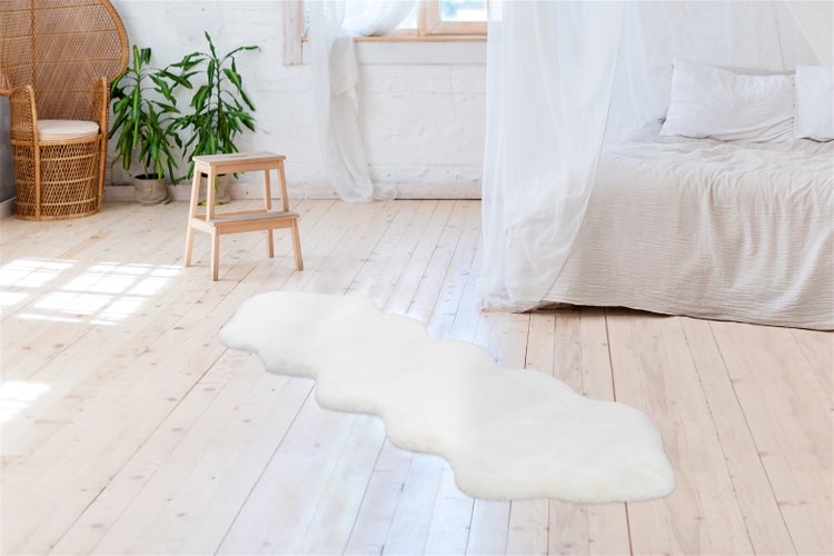 Weißer Läufer in Lammfelloptik auf hellem Holzboden in einem hellen Schlafzimmer mit Bett und Korbsessel.