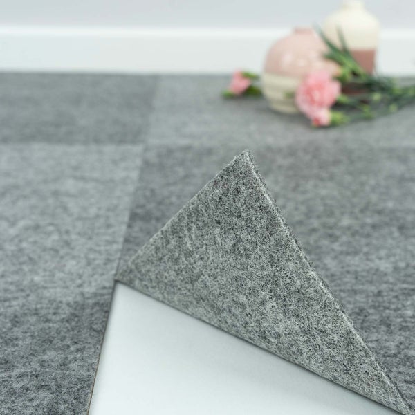 Selbstklebende Teppichfliesen in Grau mit Filzstruktur, eine Ecke ist angehoben, um die klebende Unterseite zu zeigen.
