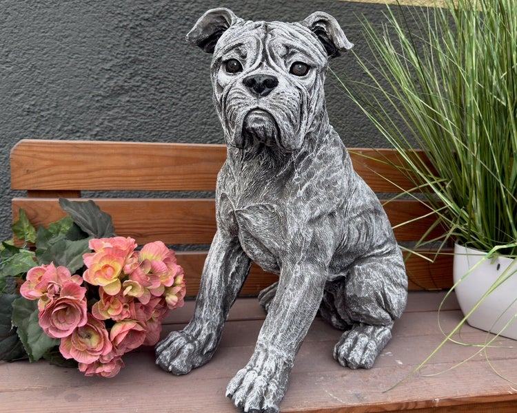 Gartenfigur in Form eines sitzenden Hundes aus Steinguss, platziert auf einer Holzbank neben Blumen.