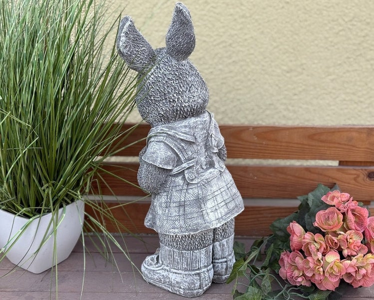 Gartenfigur Hase in Kleidung in Steinoptik von hinten, stehend auf einer Holzbank neben Pflanzen.