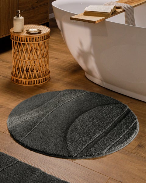 Runder, dunkelgrauer Badeteppich mit Wellenstruktur vor einer weißen Badewanne auf Holzboden, neben einem kleinen Rattantisch.