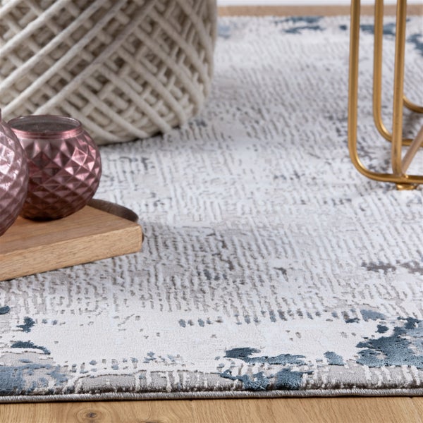 Teppich mit abstraktem Muster in Grau und Blau auf Holzboden, mit Geflechtkorb, Holztablett und Metallbein in einem hellen Raum.