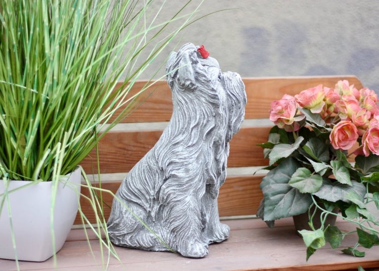 Gartenfigur Yorkshire Terrier aus Steinguss mit Schleife, sitzend auf einer Holzbank neben Topfpflanzen.
