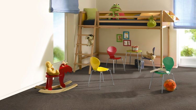 Dunkle Teppichfliesen in einem hellen Kinderzimmer mit Hochbett aus Holz, bunten Stühlen und Spielzeug.