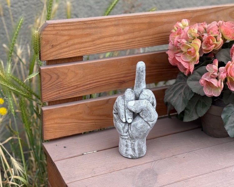 Dekorative Gartenfigur in Form einer Hand mit erhobenem Mittelfinger aus Beton auf einer Holzbank.