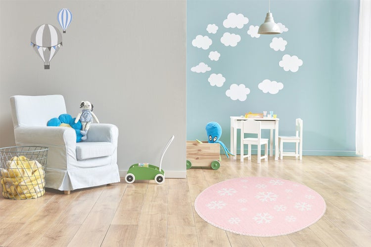Helles Kinderzimmer mit rundem rosa Teppich mit Schneeflockenmuster, Sessel, Spielzeug und Wandtattoos in Wolken- und Heißluftballonform.