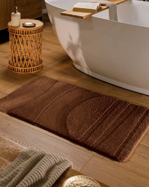 Braune Badematte mit Reliefmuster auf Holzboden vor einer weißen Badewanne.