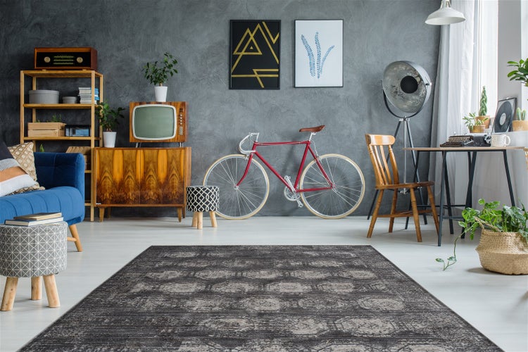 Rechteckiger Teppich mit grauem Vintage-Muster in einem Raum mit blauem Sofa, Holzmöbeln und einem Fahrrad vor einer grauen Wand.