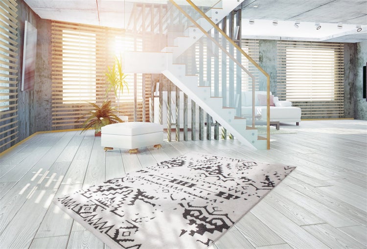 Moderner heller Wohnraum mit Holzboden, Teppich mit schwarzem geometrischem Muster, weißem Hocker und Treppe mit Glasgeländer.