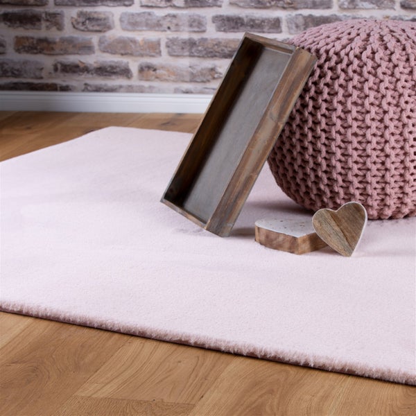Weicher Kunstfellteppich in Rosa auf Holzboden mit Holztablett, gestricktem Pouf und Dekoherzen vor einer Wand in Backsteinoptik.