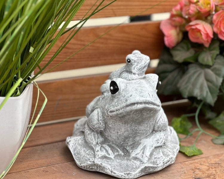 Gartenfigur Frosch aus Stein mit kleinem Frosch auf dem Rücken, dekorativ platziert vor Holz und Pflanzen.