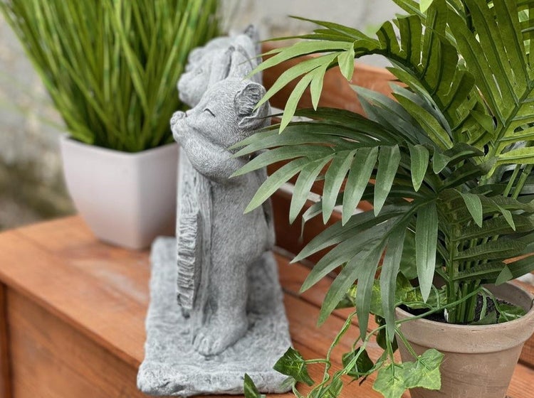 Steinfigur Katzenpaar als Gartendeko auf Holzuntergrund zwischen zwei Topfpflanzen.