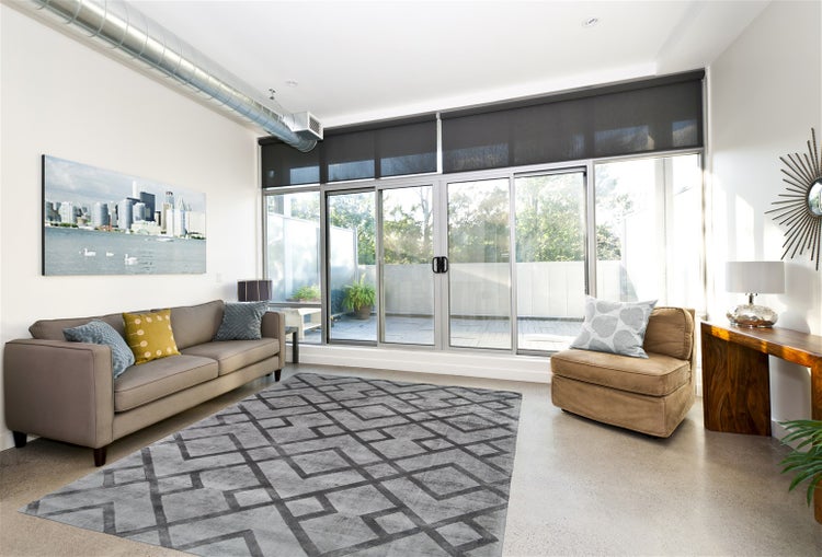 Modernes, helles Wohnzimmer mit großem grauem Teppich mit geometrischem Muster, Sofa, Sessel und bodentiefen Glasschiebetüren zum Balkon.