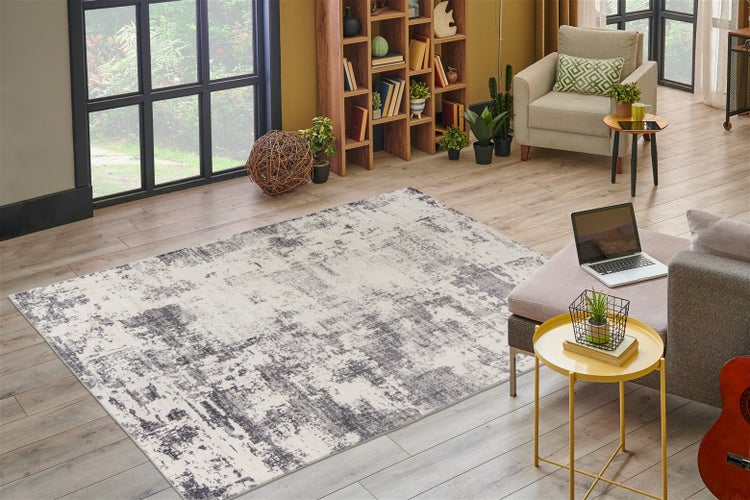 Moderner Wohnraum mit abstrakt gemustertem Teppich in Grau und Creme auf hellem Holzboden, mit Bücherregal, Sessel und großem Fenster.