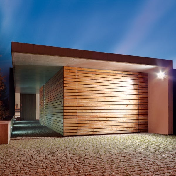 Moderne Garage mit Holzfassade und Flachdach in der Abenddämmerung mit Außenbeleuchtung und gepflasterter Einfahrt.