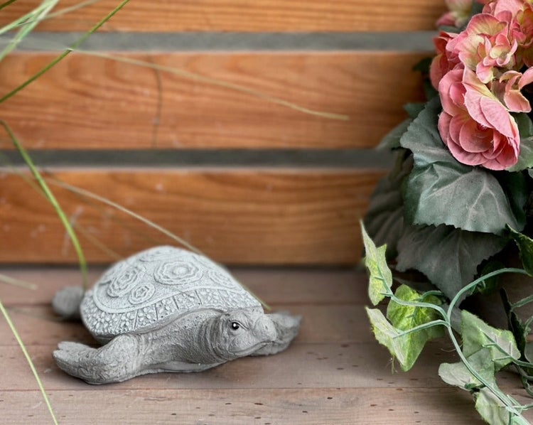 Gartenfigur in Form einer Schildkröte aus Stein auf Holzdielen neben einer Pflanze.