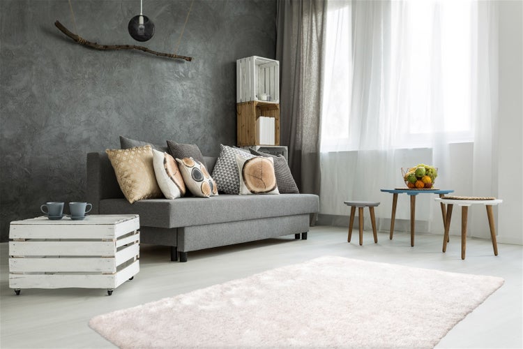 Helles Wohnzimmer mit grauem Sofa, verschiedenen Kissen, Holztischen, weißem Teppich und einer strukturierten grauen Wand.