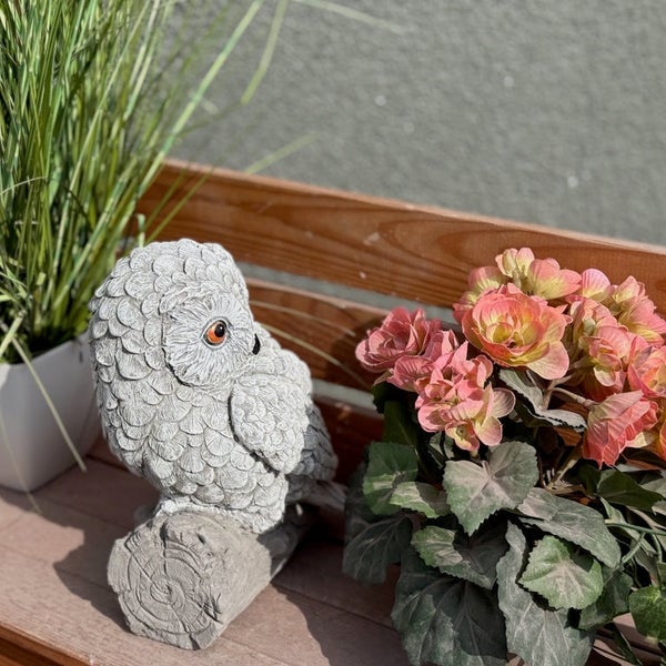 Dekorative Eulenfigur aus Stein für den Garten auf einem Baumstamm sitzend neben einer blühenden Pflanze auf einer Holzbank.