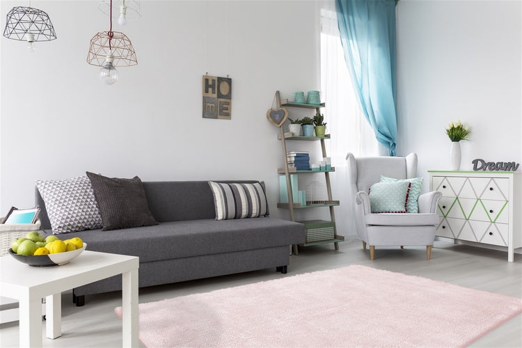 Helles Wohnzimmer mit grauem Sofa, rosa Hochflor-Teppich, hellem Ohrensessel und Kommode. Moderne Pendelleuchten und Leiterregal.