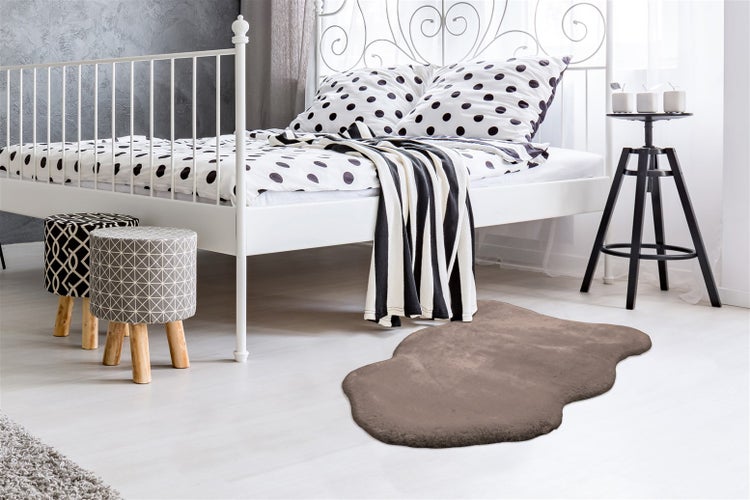 Modernes Schlafzimmer mit braunem Kunstfellteppich, weißem Metallbett und gemusterten Hockern auf hellem Boden.