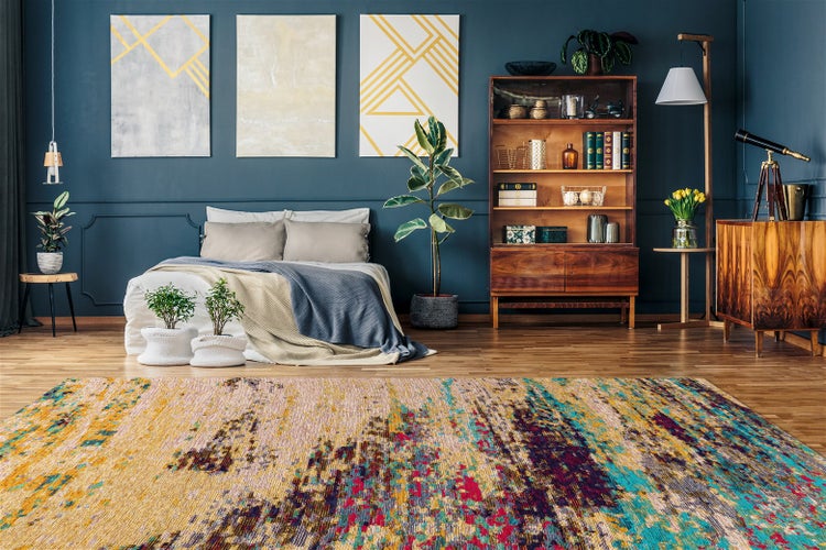 Modern eingerichtetes Schlafzimmer mit buntem, abstraktem Teppich auf Holzboden, dunkler Wand mit Bildern, Holzmöbeln und Pflanzen.