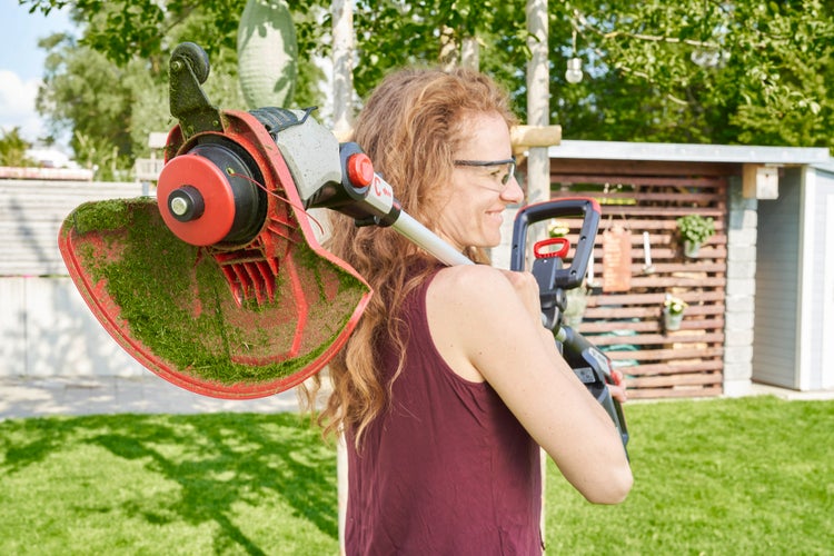 Eine Frau mit Schutzbrille trägt einen elektrischen Rasentrimmer auf der Schulter in einem Garten.