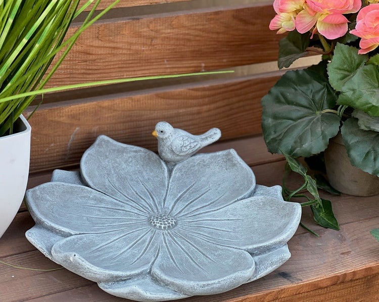 Stein-Vogeltränke in Blumenform mit dekorativer Vogelfigur auf einer Holzoberfläche mit Pflanzen