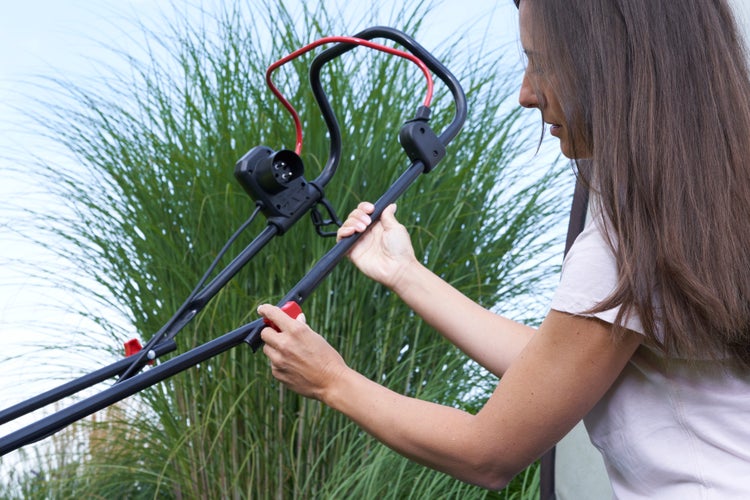 Person montiert oder verstellt den Holm eines Elektro Rasenmähers im Garten vor hohem Ziergras.