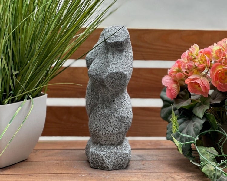 Abstrakte Gartenfigur aus grauem Stein mit strukturierter Oberfläche, platziert zwischen grünem Gras und rosa Blumen auf Holzuntergrund.