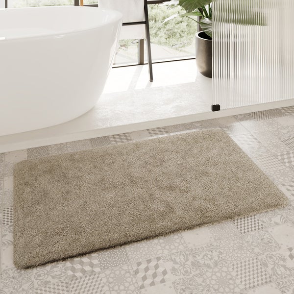 Taupefarbene Hochflor-Badematte rechteckig auf gemusterten Fliesen im hellen Badezimmer neben einer weißen freistehenden Badewanne.