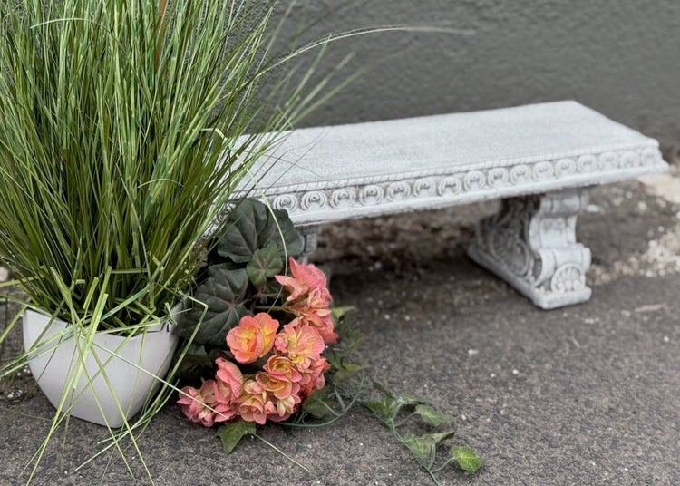 Eine kleine dekorative Gartenbank aus Stein mit Ornamenten, kombiniert mit einem Topf voller Gras und künstlichen Blumen.
