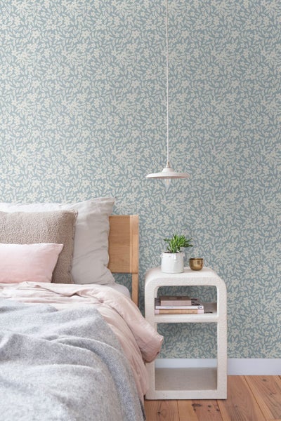 Helles Schlafzimmer mit Tapete mit floralem Muster aus Blüten und Blättern hinter einem Bett mit Holz-Kopfteil und einem Nachttisch.