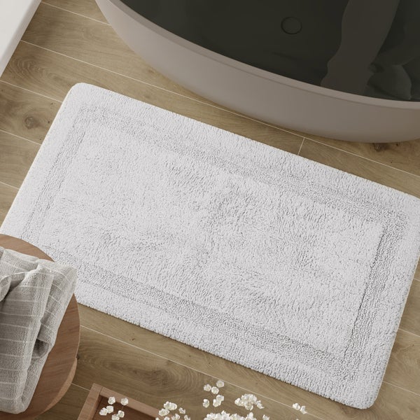 Rechteckige Badematte auf einem Boden in Holzoptik in einem hellen Badezimmer neben einer Badewanne.
