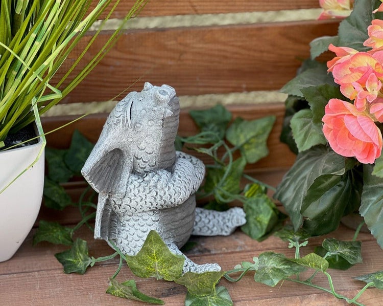 Kleine graue Drachenfigur in Steinoptik, sitzend mit verschränkten Armen und nach oben blickend, dekoriert mit künstlichem Efeu auf Holz.