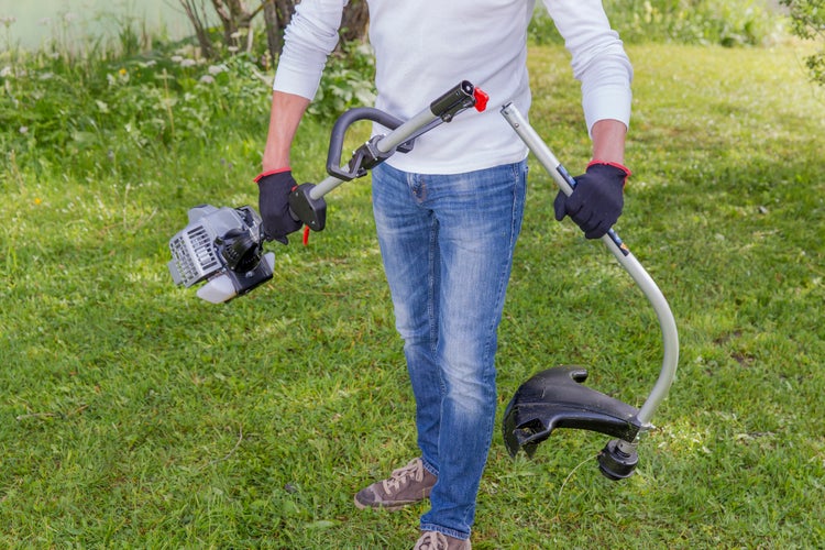 Mann bei der Gartenarbeit mit einem Benzin Rasentrimmer auf einer Wiese vor einer Berglandschaft.