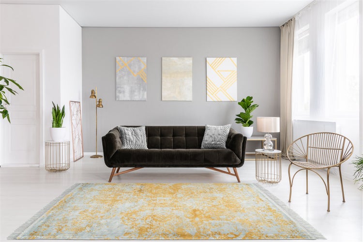 Modernes Wohnzimmer mit dunkelbraunem Samtsofa, gelb-blau gemustertem Teppich, goldfarbenem Metallstuhl und abstrakten Bildern an der Wand.