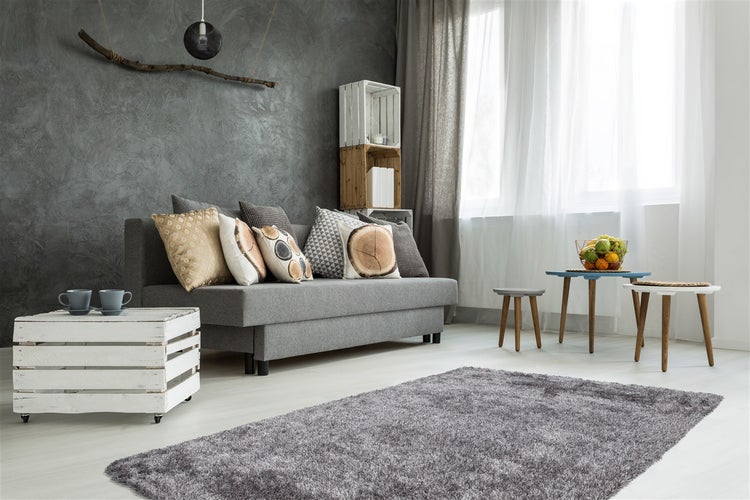 Modernes Wohnzimmer mit grauem Sofa, strukturierten Kissen, einem Hochflor-Teppich und Couchtischen aus Holzkisten vor einer dunklen Akzentwand.