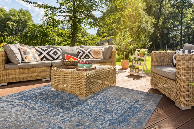 Moderne Terrasse mit blau-grau gemustertem Außenteppich, Rattan-Ecksofa, passendem Couchtisch und Sessel auf einem Holzdeck im grünen Garten.