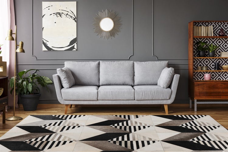 Modernes Wohnzimmer mit grauem Dreisitzer-Sofa, geometrischem Teppich, Holzregal und goldener Stehlampe vor dunkelgrauer Wand mit Zierleisten.
