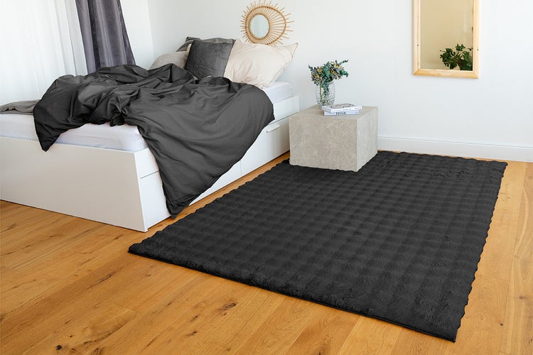 Moderner anthrazitfarbener Strukturteppich auf Holzboden in einem hellen, aufgeräumten Schlafzimmer mit weißem Bett und Steinnachttisch.