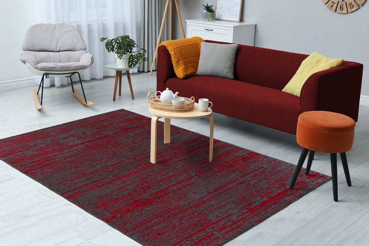 Modernes Wohnzimmer mit einem rot-grau gemusterten Teppich auf hellem Boden, rotem Sofa, grauem Schaukelstuhl und kleinem Holztisch.