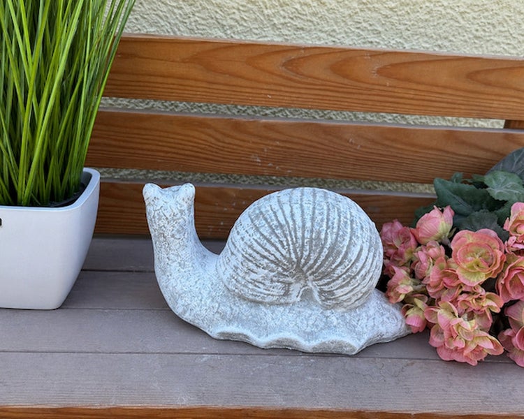 Dekorative Schneckenfigur aus Stein für den Garten auf einer Holzbank neben einer Pflanze und Blumen.