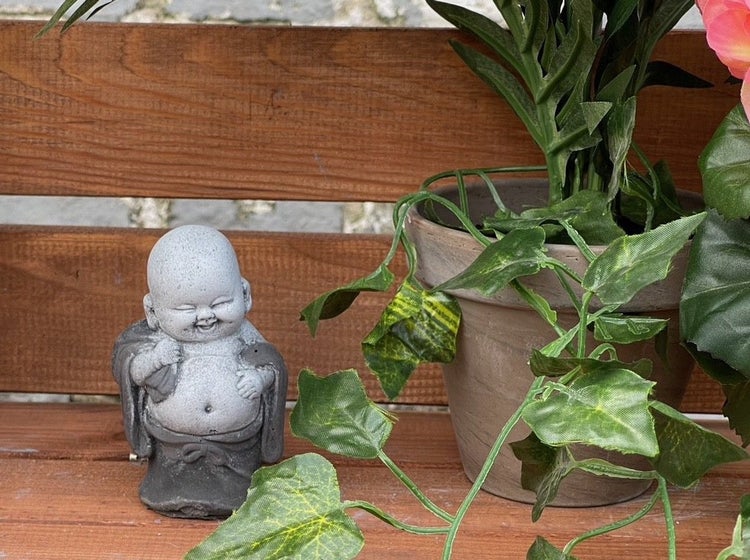 Kleine lachende Buddha Gartenfigur aus Stein auf einer Holzbank neben einem Blumentopf mit künstlicher Pflanze.