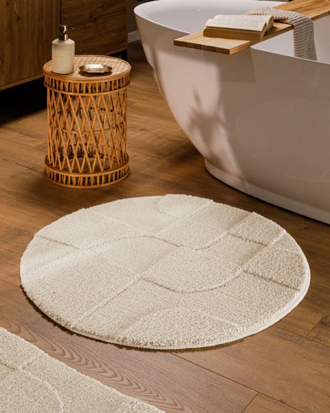 Runder Badevorleger mit Strukturmuster auf Holzboden neben einer weißen freistehenden Badewanne und einem Rattan Beistelltisch.